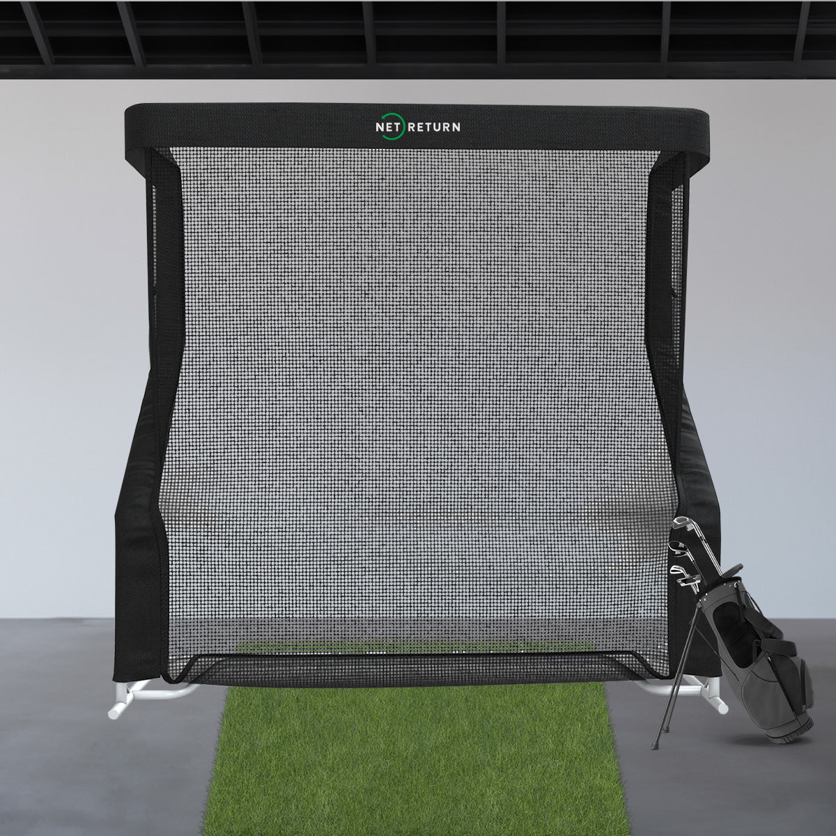 A golf practice net with a grass mat in front and a golf bag with clubs to the side, set in an indoor space.